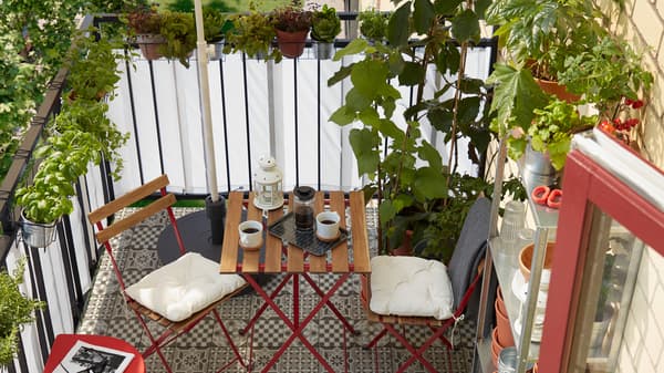 Un pequeño balcón con muchas hierbas y plantas, una estantería con macetas, un parasol, dos sillas y una mesa.