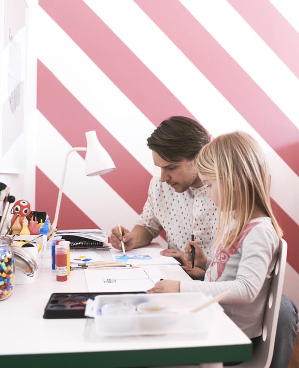 Un père et sa fille assis à un bureau, en train de peindre, sur fond de papier peint rayé.