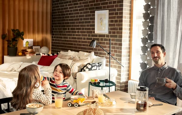 Un père prend son petit déjeuner, installé à table; il a le visage serein, tandis que ses enfants jouent avec de la nourriture à côté de lui.