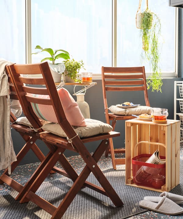 Un petit coin café sur un balcon ensoleillé formé par des chaises pliantes et une caisse en bois KNAGGLIG placée verticalement faisant office de petite table.