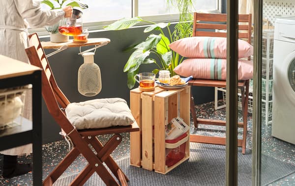 Un petit coin café sur un balcon ensoleillé formé par des chaises pliantes et une caisse en bois KNAGGLIG faisant office de petite table placée au milieu.