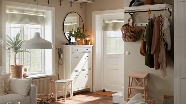 Un petit hall d’entrée avec une porte blanche, une armoire à chaussures blanche, un miroir rond, une étagère à chapeaux blanche où sont suspendus des manteaux.