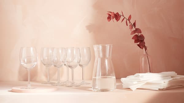 Un piano rosa antico con bicchieri e una caraffa della serie SVALKA . Accanto, un ramo fiorito e dei tovaglioli. 