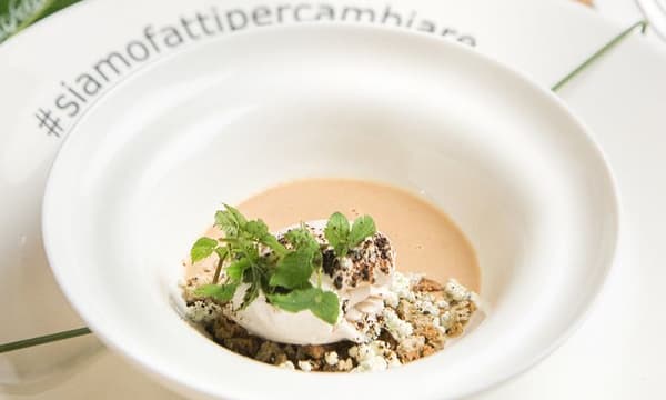 Un piatto bianco con gelato fatto con SILL MATJES aringhe, burro di frutta secca selvatica mista, foglie di aegopodium podagraria, pane al lichene islandico essiccato e sbriciolato e terra di radici fermentate.
