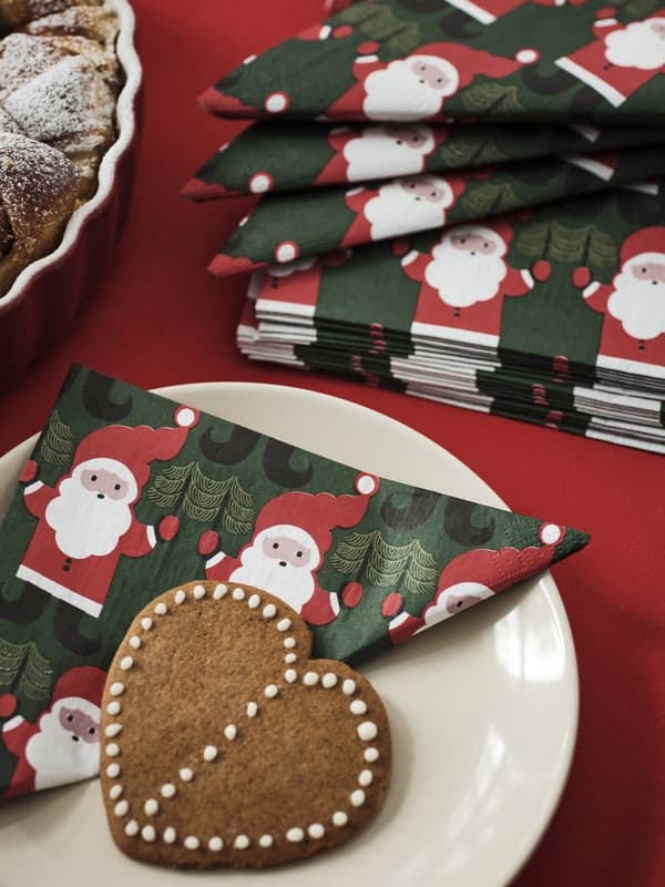 Un piatto con un biscotto a forma di cuore decorato con ghiaccia bianca e un tovagliolo con Babbo Natale verde, bianco e rosso, ripiegato. A lato, si intravede una torta in teglia.