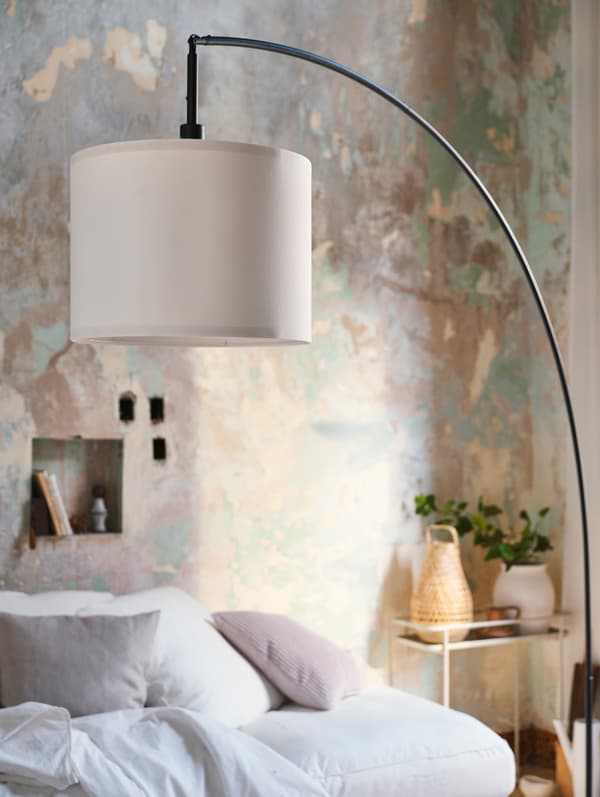 Un pied de lampadaire voûté noir avec abat-jour blanc. Un support de plante de couleur crème se trouve en arrière-plan.