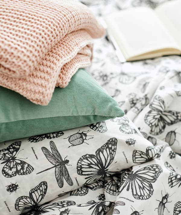 Un plaid en tricot rose et un coussin en velours vert placés au-dessus d’une couette et d’un oreiller aux housses assorties à motifs d’insectes.