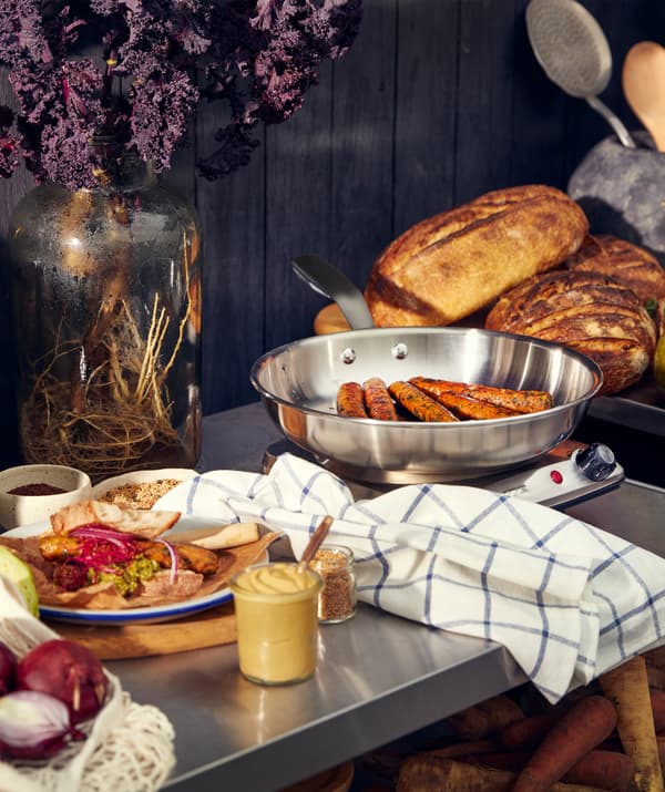 Un plan de travail métallique avec une assiette remplie de nourriture, des tranches de pain et des saucisses sur une balance.
