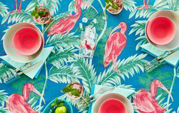 Un plateau de table avec une nappe en tissu aux motifs tropicaux colorés en rose, vert et bleu. Une vaisselle assortie est posée sur la table.