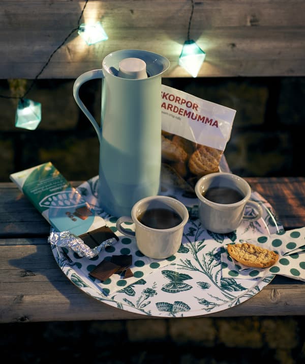 Un plateau MUSTIGHET est placé sur un banc dans un parc en pleine soirée. Sur celui-ci on retrouve un pichet isotherme BEHÖVD, des tasses à café, des petits pains croquants et du chocolat.