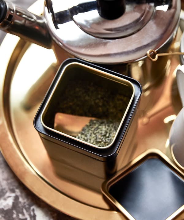 Un plateau rond doré avec un kit pour le thé: une bouilloire, une boîte avec son couvercle enlevé révélant du thé vert à l’intérieur, des tasses sur le côté.