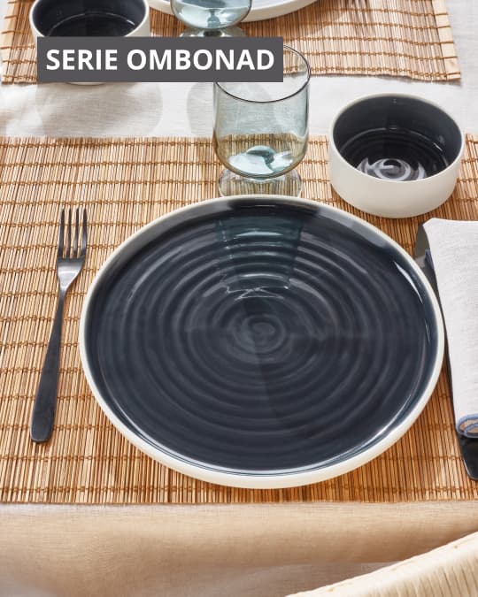Un plato grande negro con bordes blancos de la serie OMBONAD sobre un individual de mimbre, cristalería y cubiertos.