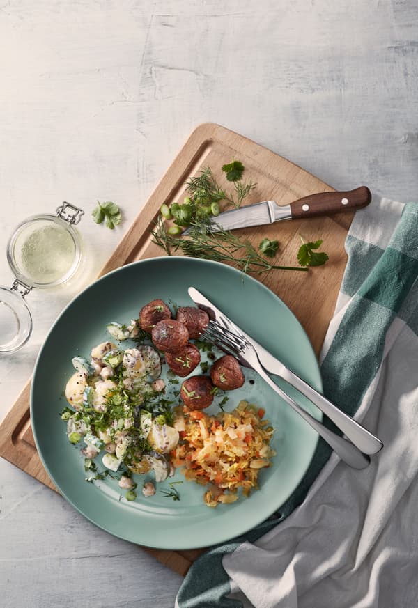 Un plato turquesa con albondigas vegetales, ensalada de papas y kimchi, todo sobre una tabla para cortar con un cuchillo, hierbas y paños de cocina.