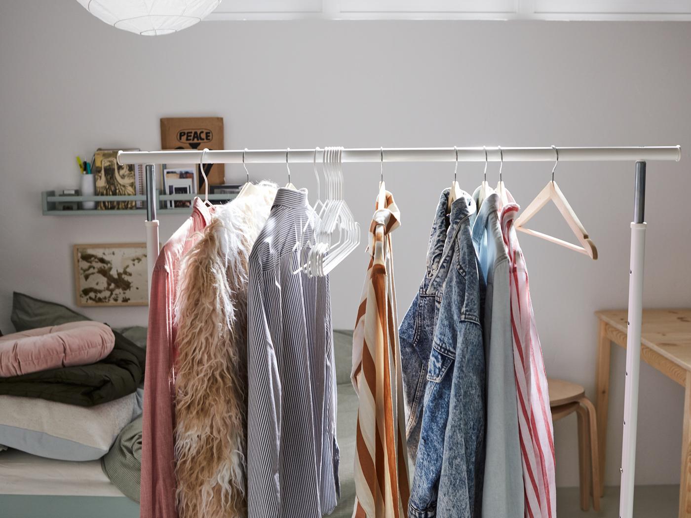 Un portant RIGGA blanc avec des vêtements accrochés sur des cintres à côté d’une table en pin et d’un lit où plusieurs coussins et du linge de lit sont entassés.