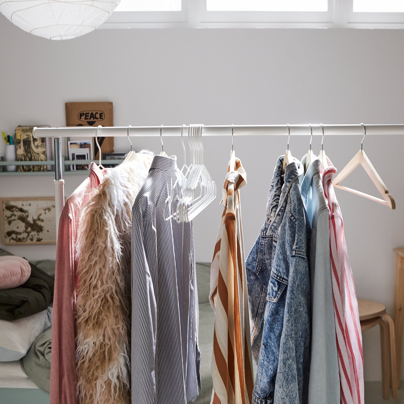 Un portant RIGGA blanc avec des vêtements sur des cintres à côté d’une table en pin et d’un lit avec une pile de coussins et de la literie.