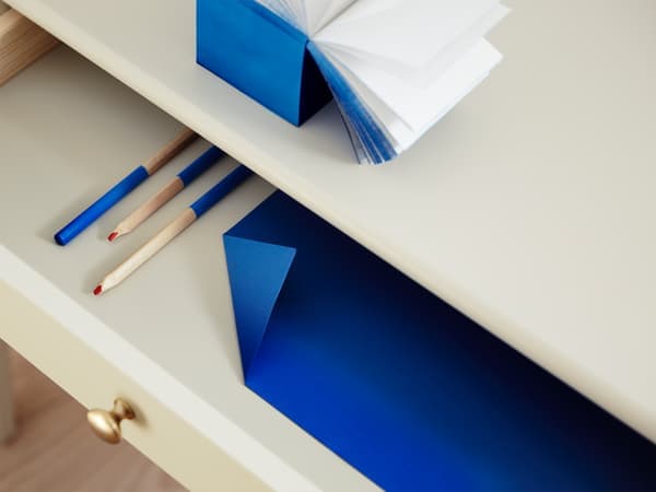Un primer pla d’un calaix obert amb llapis de colors i un sobre blau a dins; a sobre, un quadern obert de tapa blava.