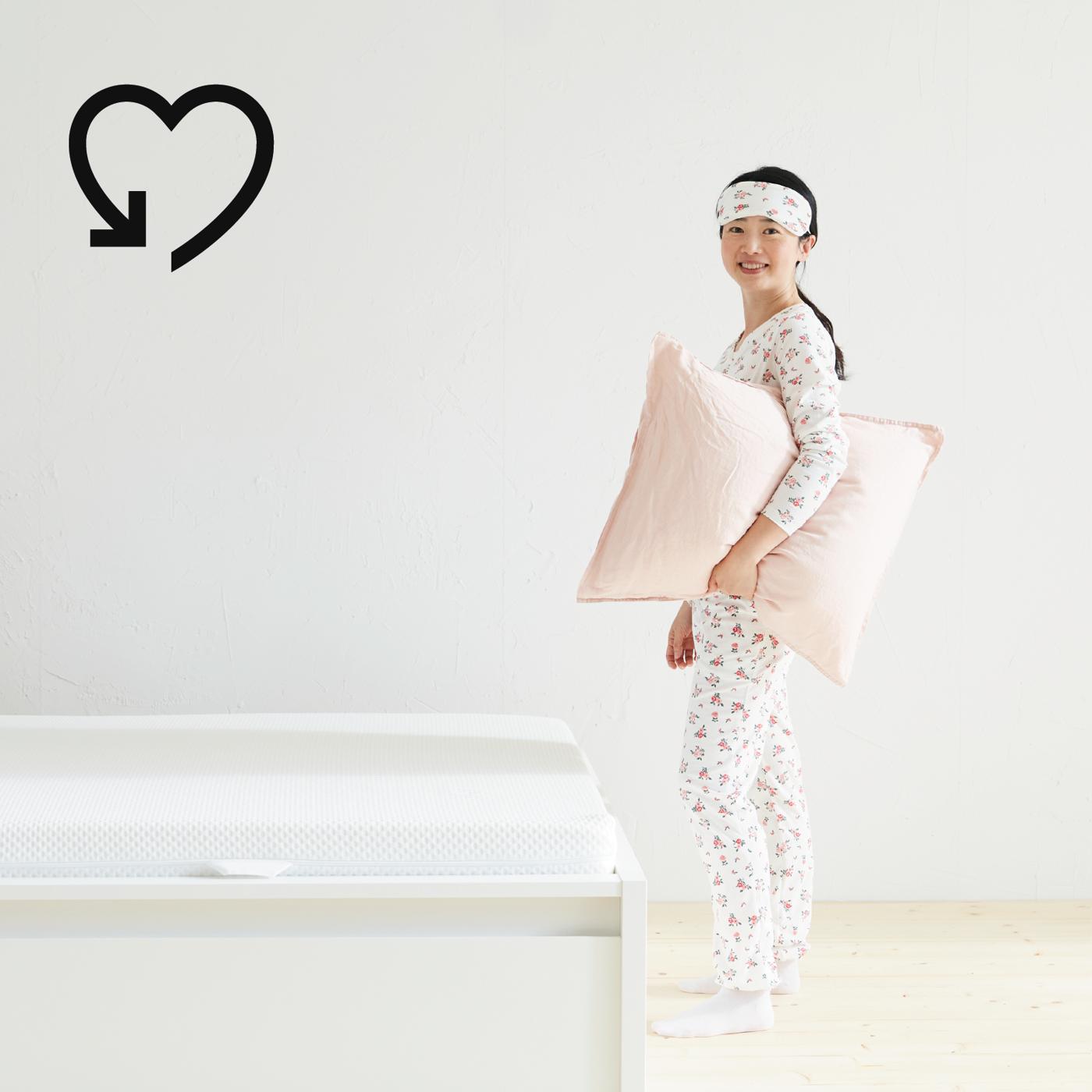 Un ragazza in pigiama con un cuscino tra le braccia è accanto ad un letto MALM bianco con materasso.