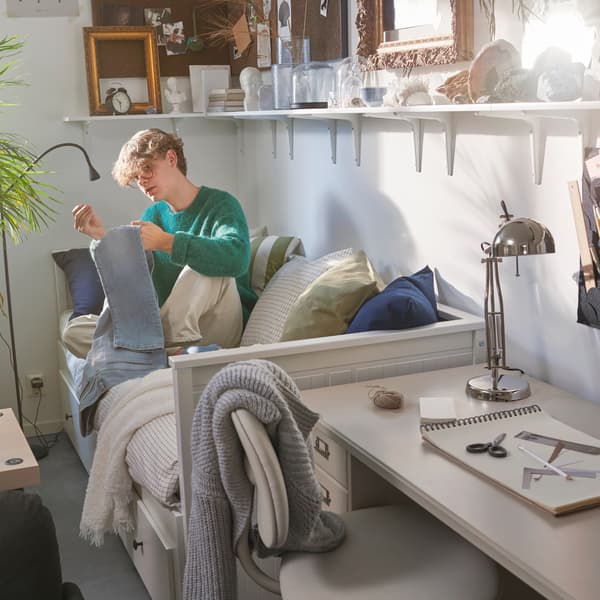 Un ragazzo seduto su un letto divano HEMNES bianco, con accanto una scrivania beige con una lampada da lavoro nichelata e una sedia girevole beige.