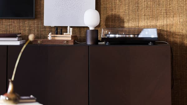 Un rangement BESTÅ brun foncé contre un mur en jute, une lampe de table BLEKKLINT et un cadre avec illustration SYMFONISK avec une enceinte Wi-Fi.
