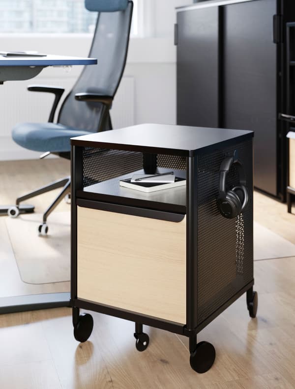 Un rangement mobile BEKANT noir sur lequel sont posés un carnet et un casque, devant une chaise de bureau bleue JÄRVFJÄLLET.