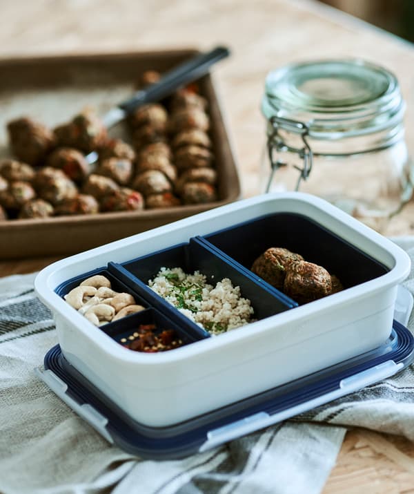 Un récipient en plastique blanc avec des inserts bleus, chacun contenant des aliments différents, posé sur un torchon de cuisine en lin à côté d’un plateau rempli de boulettes végétariennes.
