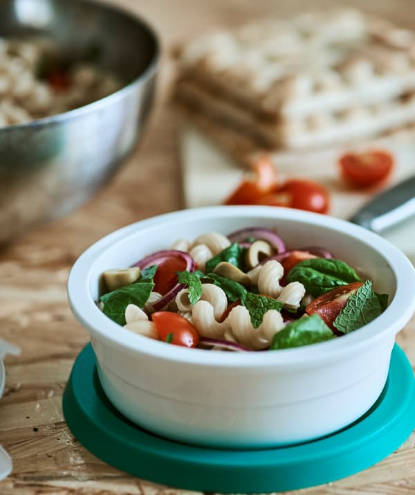 Un récipient rond en plastique blanc contenant de la tomate, des oignons rouges et une salade de pâtes aux olives vertes, sur laquelle se trouve un couvercle bleu, le tout posé sur une table en bois.