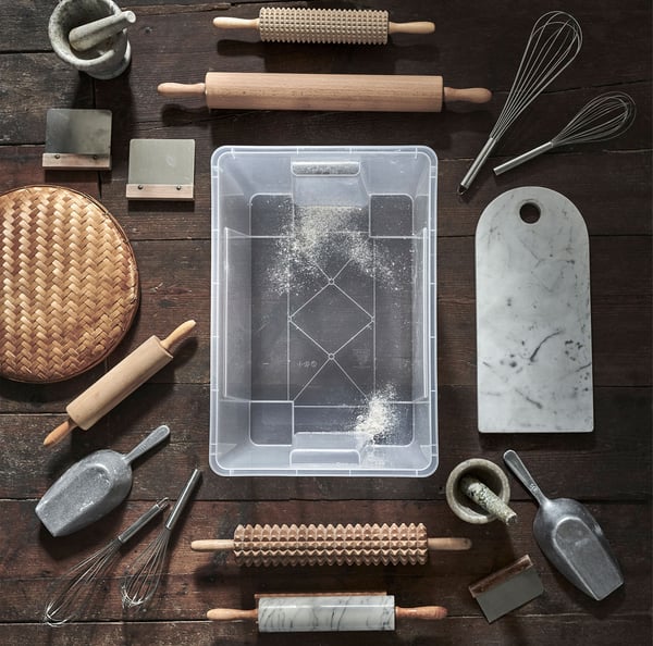 Un recipient SAMLA transparent așezat pe o podea din lemn, înconjurat de diverse accesorii pentru copt.