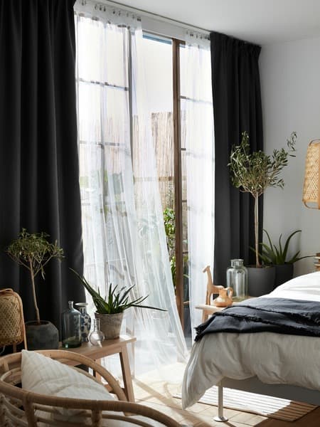 Un rideau filet blanc LILL flotte au vent devant une fenêtre ouverte dans une chambre à coucher par une journée ensoleillée. Il touche presque le lit.