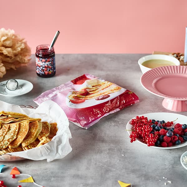Un sac de crêpes provenant de l’Épicerie suédoise IKEA est placé au centre d’une table grise. Il est entouré de confiture et de baies.