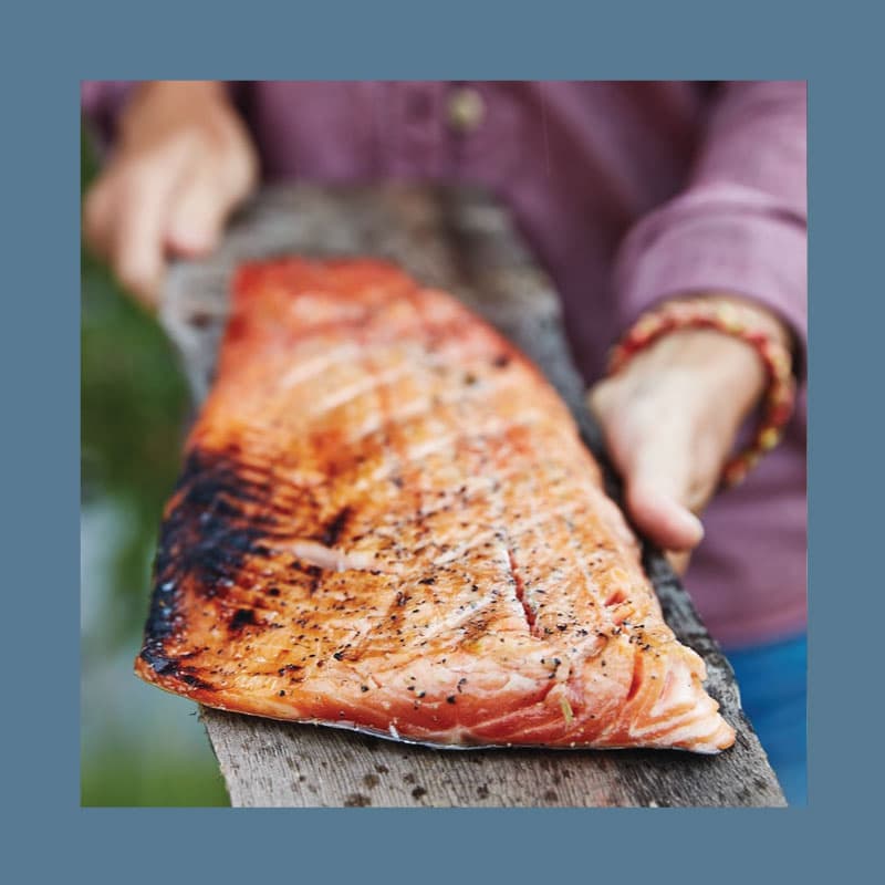 Un salmone cotto alla griglia presentato su un vassoio di corteccia da due mani.