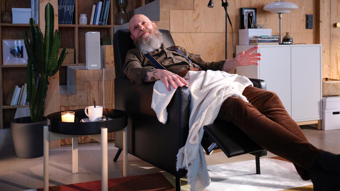 Un salon avec un homme assis dans un fauteuil inclinable GISTAD noir qui écoute de la musique à côté d’une table basse sur laquelle est posée une tasse de thé.