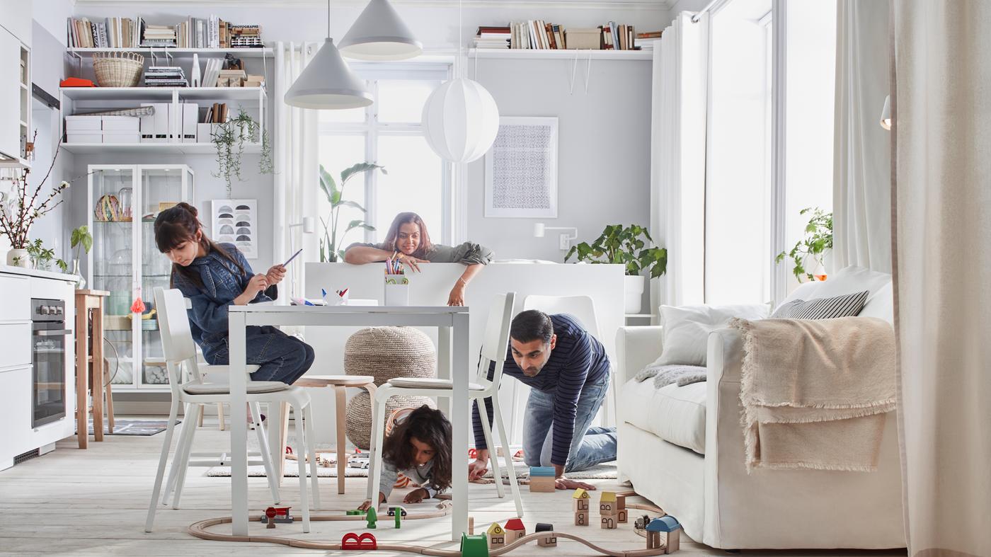 Un salón blanco y luminoso con una familia de cuatro personas jugando con un circuito de tren de madera LILLABO dispuesto por el suelo.
