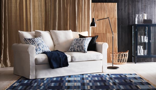 Un salón con un sofá en blanco y una alfombra azul y gris grande, una lámpara de pie, una cesta y una vitrina negra al fondo de IKEA