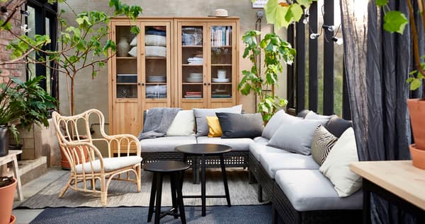 Un salón exterior con muebles modulares y un sillón de mimbre, alfombras, plantas y dos mesas de centro de IKEA.