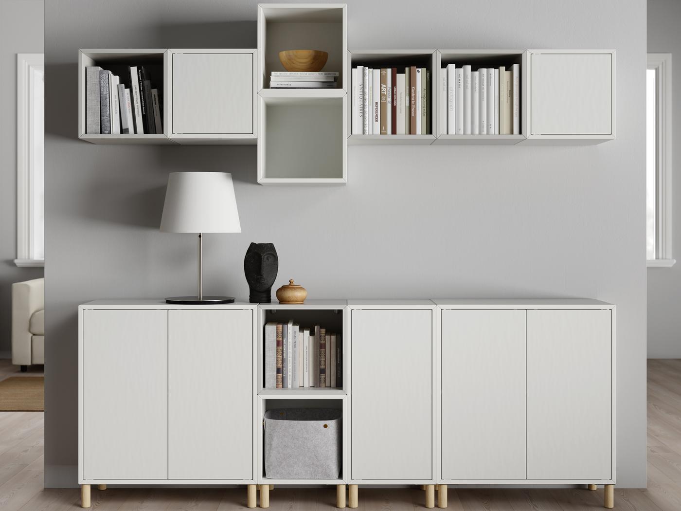 Un salon lumineux où des rangées d’armoires EKET blanc et gris clair sont fixées à un mur près du plancher.