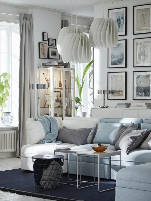 Un salotto che combina con un divano azzurro, cuscini grigi, bianchi e azzurri con 3 tavolini e un tappeto blu. Sulle pareti sono appoggiati diversi quadri, una credenza e 3 lampadari. 