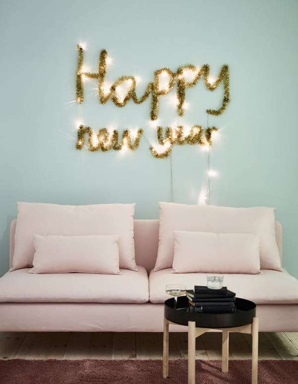 Un salotto con un divano rosa dove sulla parete l'illuminazione a LED SÄRDAL a batterie e una ghirlanda color oro che creano la scritta 