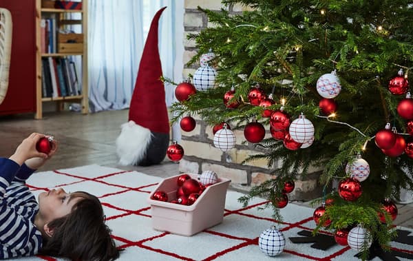 Un sapin de Noël chargé de décorations ; un garçonnet couché sur le dos à côté, sur un tapis, en train d’examiner une décoration.