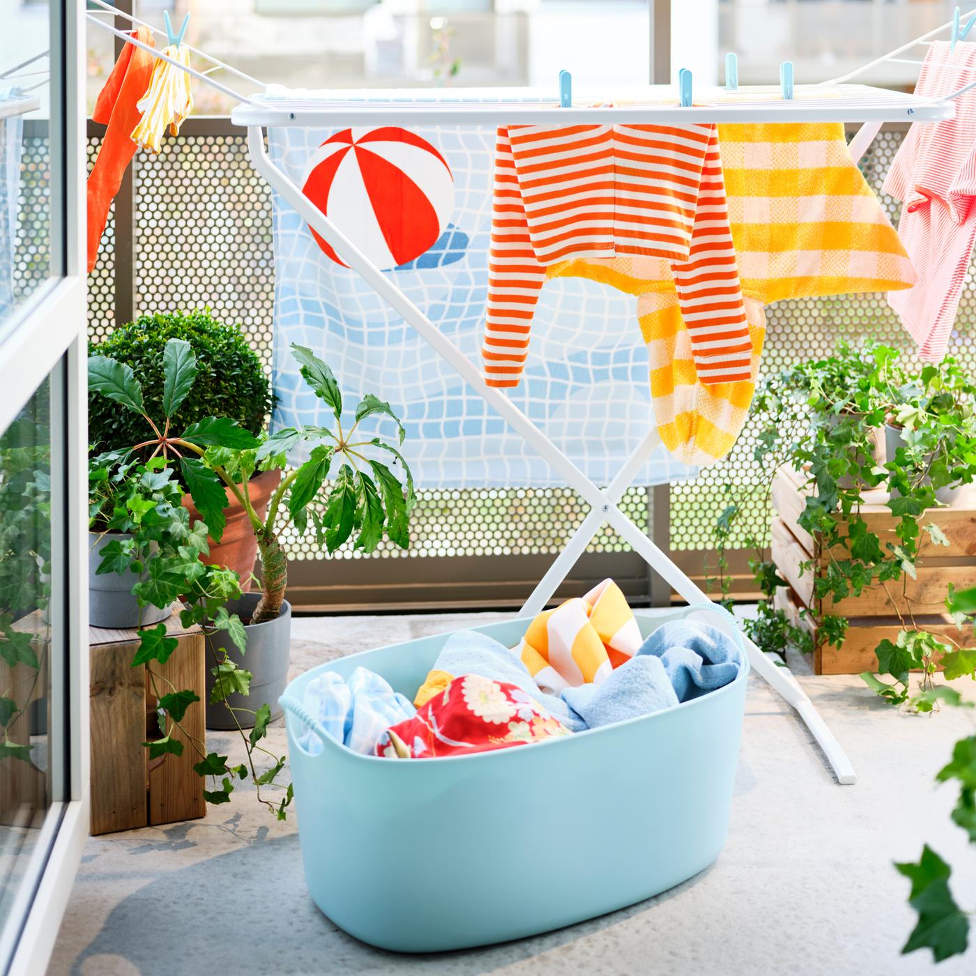 Un séchoir blanc sur un balcon avec un drap de bain et divers vêtements, plus un panier à linge bleu clair et des plantes en pot.