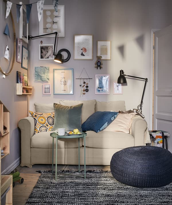 Un séjour avec un canapé deux places, un pouf bleu foncé, une petite table plateau grise et des lampes noires de chaque côté.