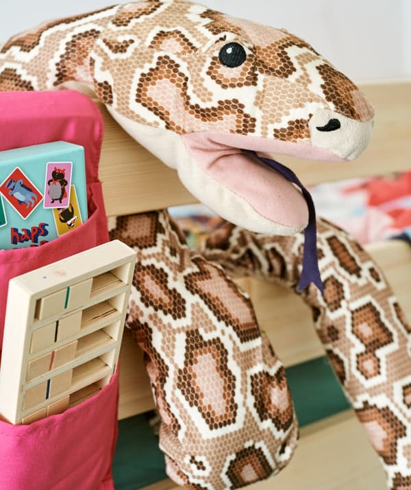Un serpent en peluche enroulé autour du cadre d’un lit en bois, à côté d’une poche de rangement en tissu contenant des jeux.