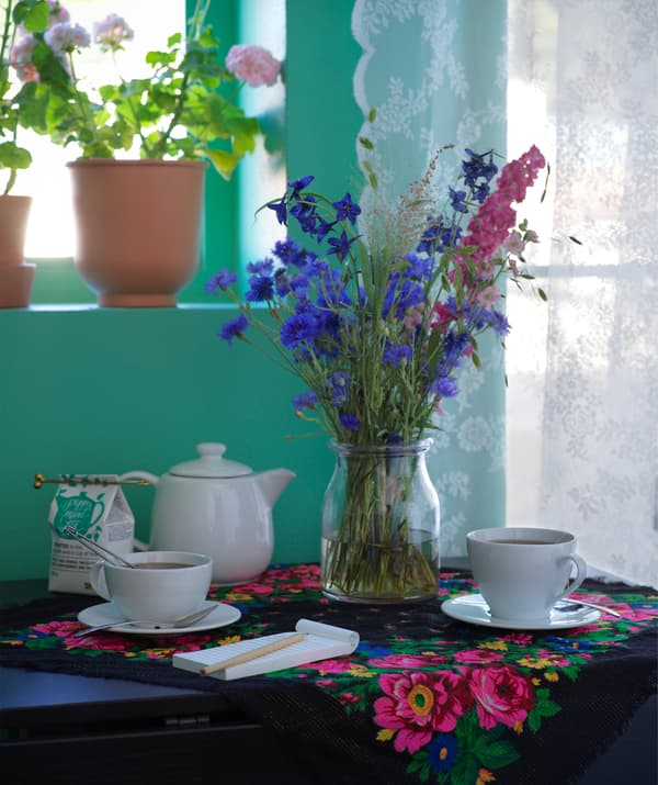 Un servizio da te con accanto un vaso di vetro con fiori colorati. C'è anche una busta di tè e un taccuino con una matita in primo piano. Tutti gli oggetti poggiano su una tovaglietta a fiori.