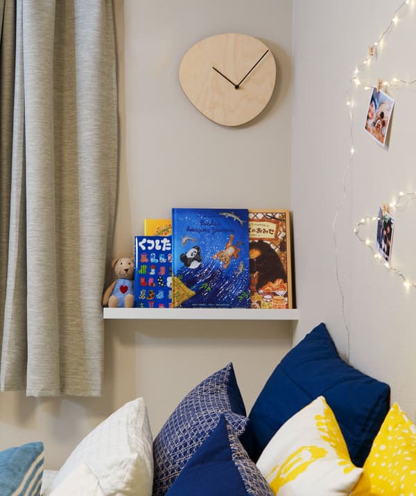 Un sinxelo reloxo de madeira montado nunha parede e un estante ateigado de libros para nenos sobre unha cama chea de coxíns.