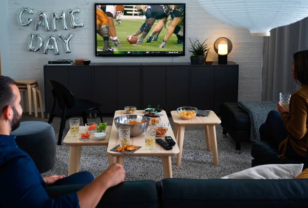 Un soggiorno con alcune persone sedute mentre stanno guardando una partita di rugby in tv. Davanti a loro, dei tavolini con cibi e alimenti.