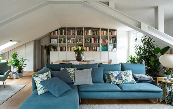 Un soggiorno con divano azzurro angolare in primo piano e tavolo e libreria di fondo.
