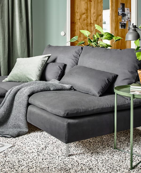 Un soggiorno con elementi grigi della serie SÖDERHAMN e un tappeto beige. Sopra il divano, alcuni cuscini e plaid. Accanto, un tavolino verde GLADOM.