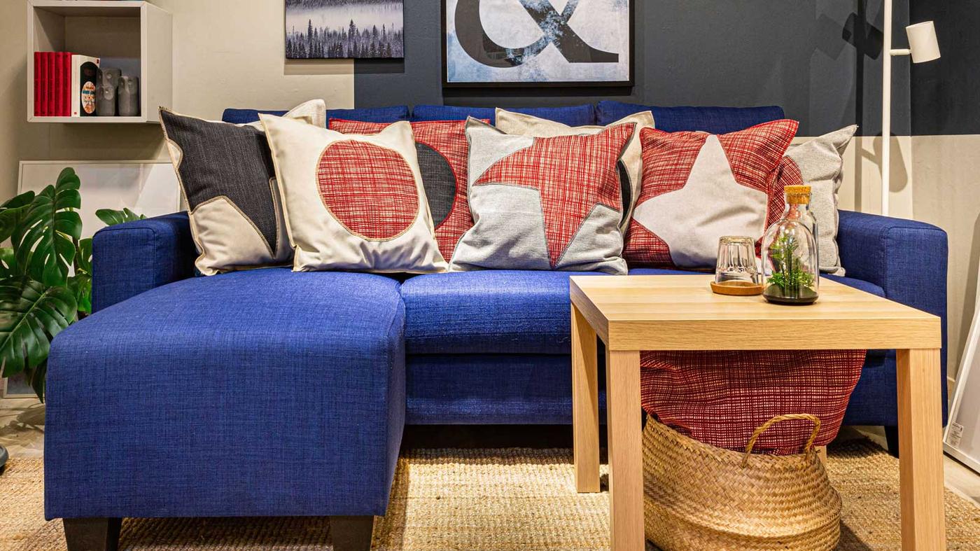 Un soggiorno con un divano/chaise lounge, un tavolino, una lampada da terra a 3 faretti NYMÅNE, una pianta in vaso all'angolo, un mobile da parete EKET e un cestino FLÅDIS.