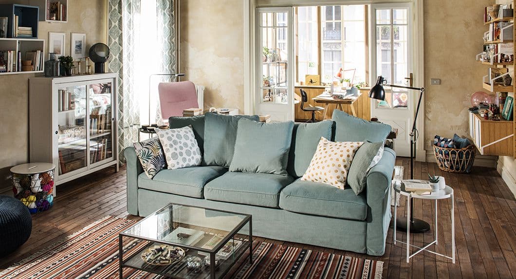 Un soggiorno con un divano verde chiaro con cuscini. Di fronte, un tavolino in vetro e metallo, e accanto un tavolino GLADOM. Sulla sinistra una vetrina, un tavolino, un contenitore e una poltrona rosa VEDBO. Sulla destra una scaffalatura e una cesta di rattan.