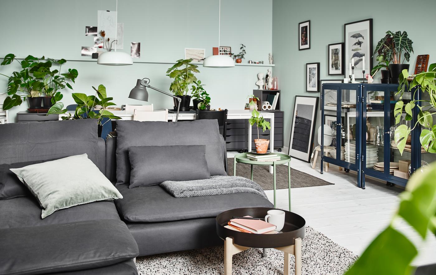 Un soggiorno ideale secondo la interior designer Therese Ericsson: ci sono un divano grigio della serie di divani SÖDERHAMN con cuscini, pareti verde chiaro, piante in vaso e diversi mobili con vetrina.