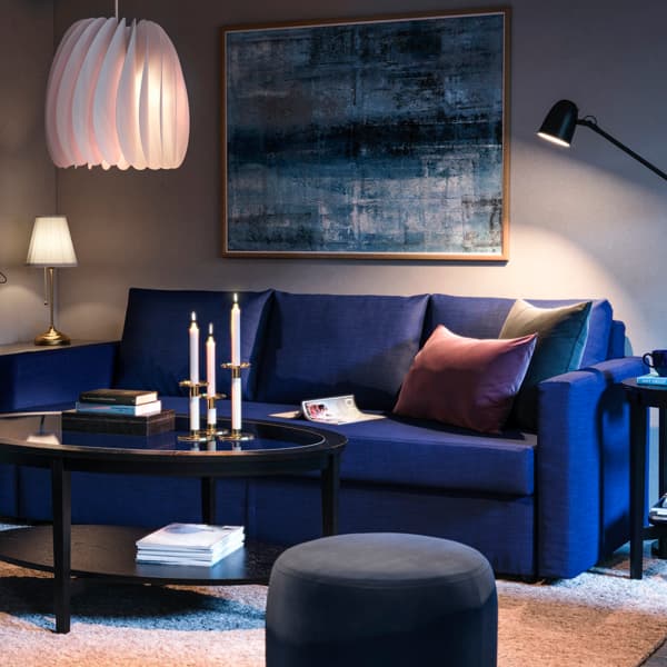 Un soggiorno illuminato da una lampada a sospensione SKYMNINGEN e da una ÅRSTID. Nel soggiorno ci sono un tavolino con un divano blu, un tavolino nero con delle candele e libri. Alla parete, un quadro.